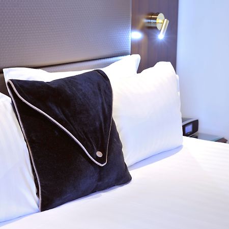 Отель Stamford Plaza Мельбурн Экстерьер фото The photo shows a neatly arranged bed with white linens. There are several pillows, with one black pillow featuring a distinctive design on top of the white pillows. The headboard of the bed is visible, along with a stylish wall-mounted light fixture