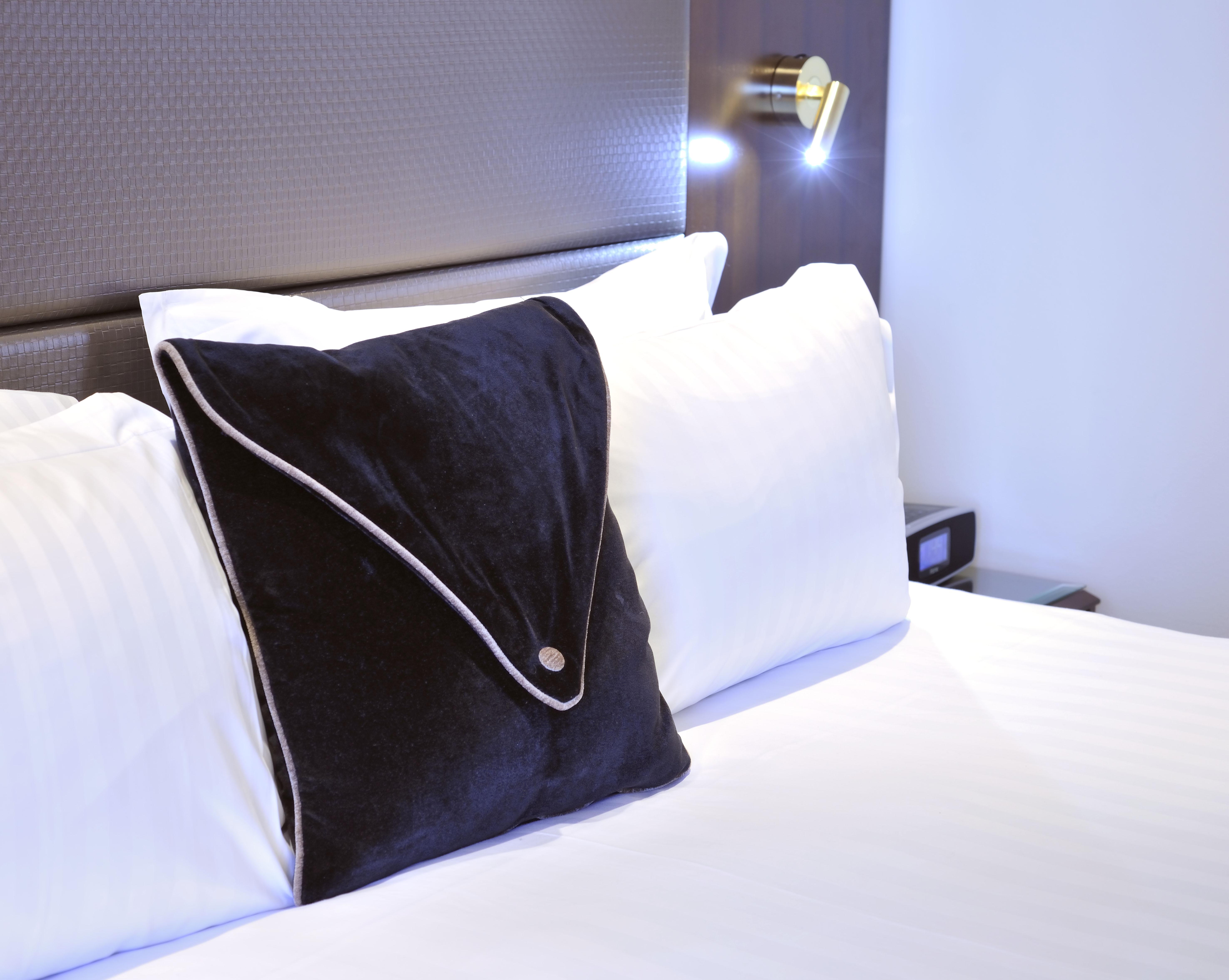 Отель Stamford Plaza Мельбурн Экстерьер фото The photo shows a neatly arranged bed with white linens. There are several pillows, with one black pillow featuring a distinctive design on top of the white pillows. The headboard of the bed is visible, along with a stylish wall-mounted light fixture