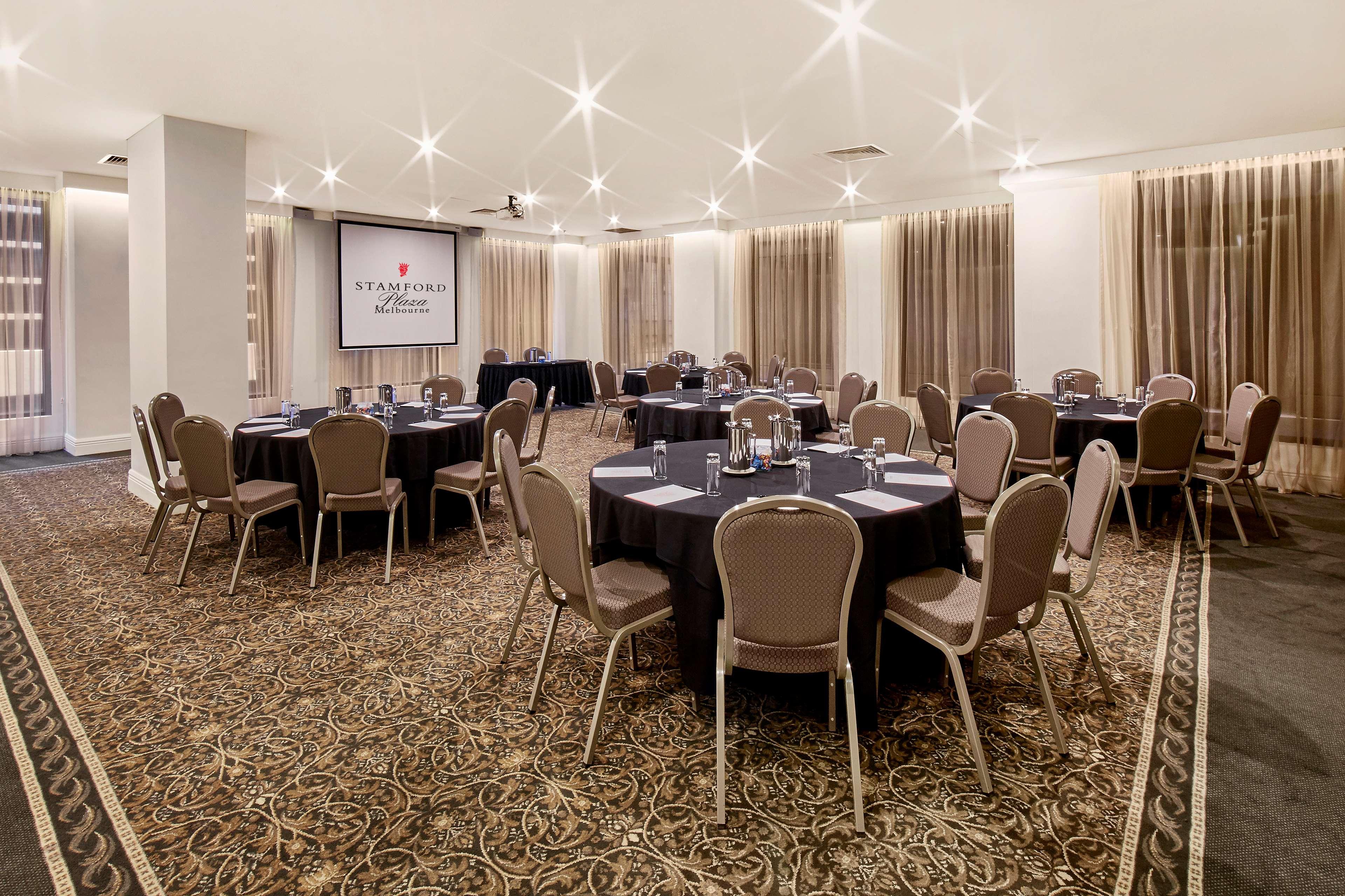 Отель Stamford Plaza Мельбурн Экстерьер фото The photo shows a well-lit event space set up for a gathering or meeting. There are several round tables covered with black tablecloths, each surrounded by beige chairs. In the background, a large screen displays a logo or branding, likely related to