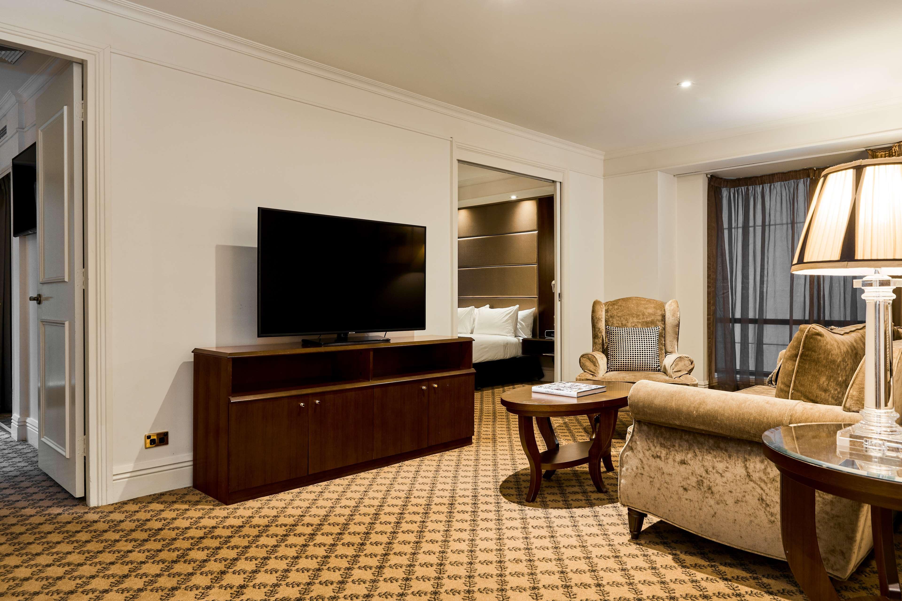 Отель Stamford Plaza Мельбурн Экстерьер фото The photo shows a well-furnished room, likely a hotel suite or a living area. There is a large flat-screen television mounted on a wooden entertainment unit. In front of the TV, there is a round wooden coffee table. To the side, a comfortable armchai