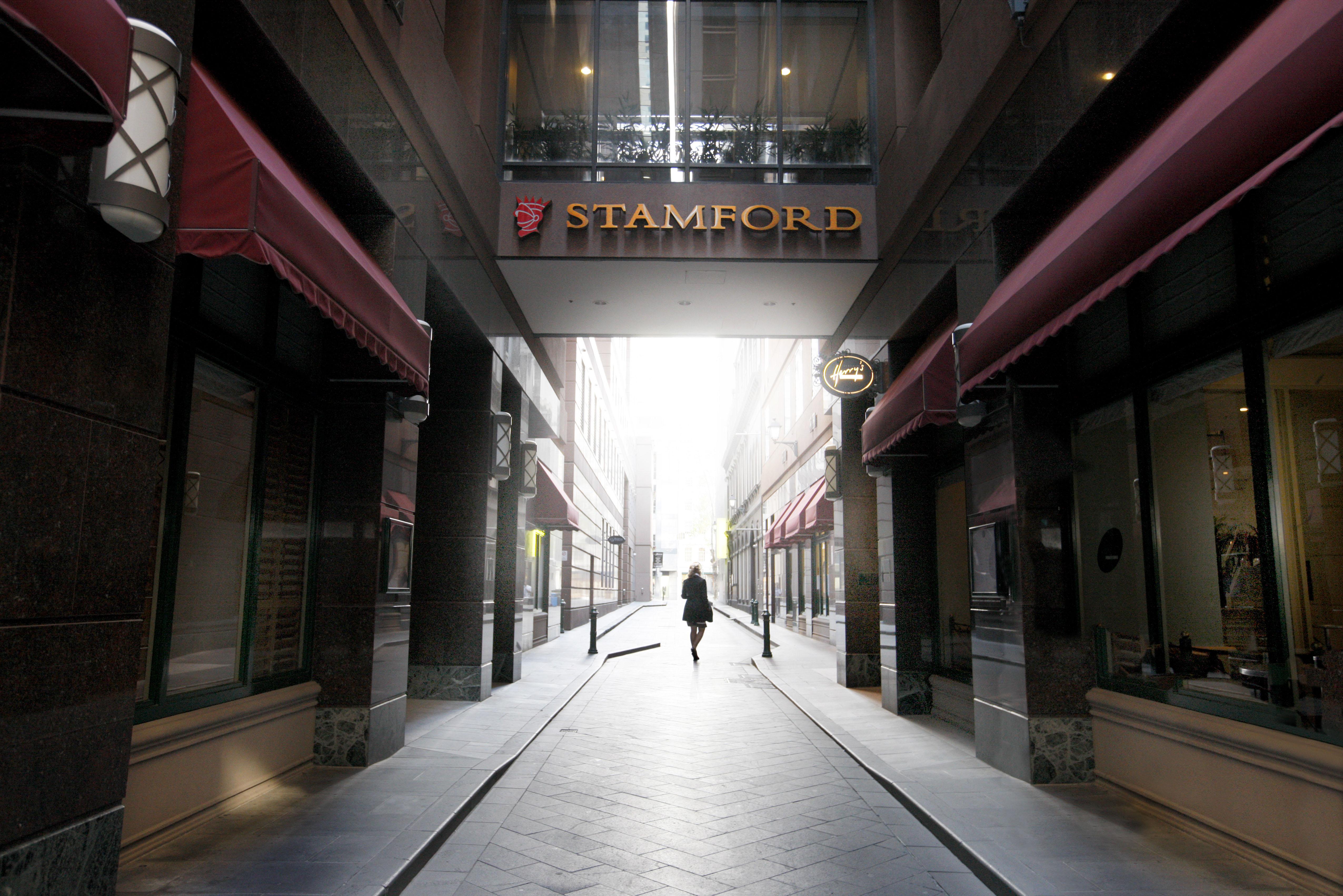 Отель Stamford Plaza Мельбурн Экстерьер фото The photo depicts a narrow alley or passageway, possibly in an urban setting. On one side, there are storefronts with canopies, and on the opposite side, there is a prominent sign reading "STAMFORD." The pathway is lined with buildings, creating a se