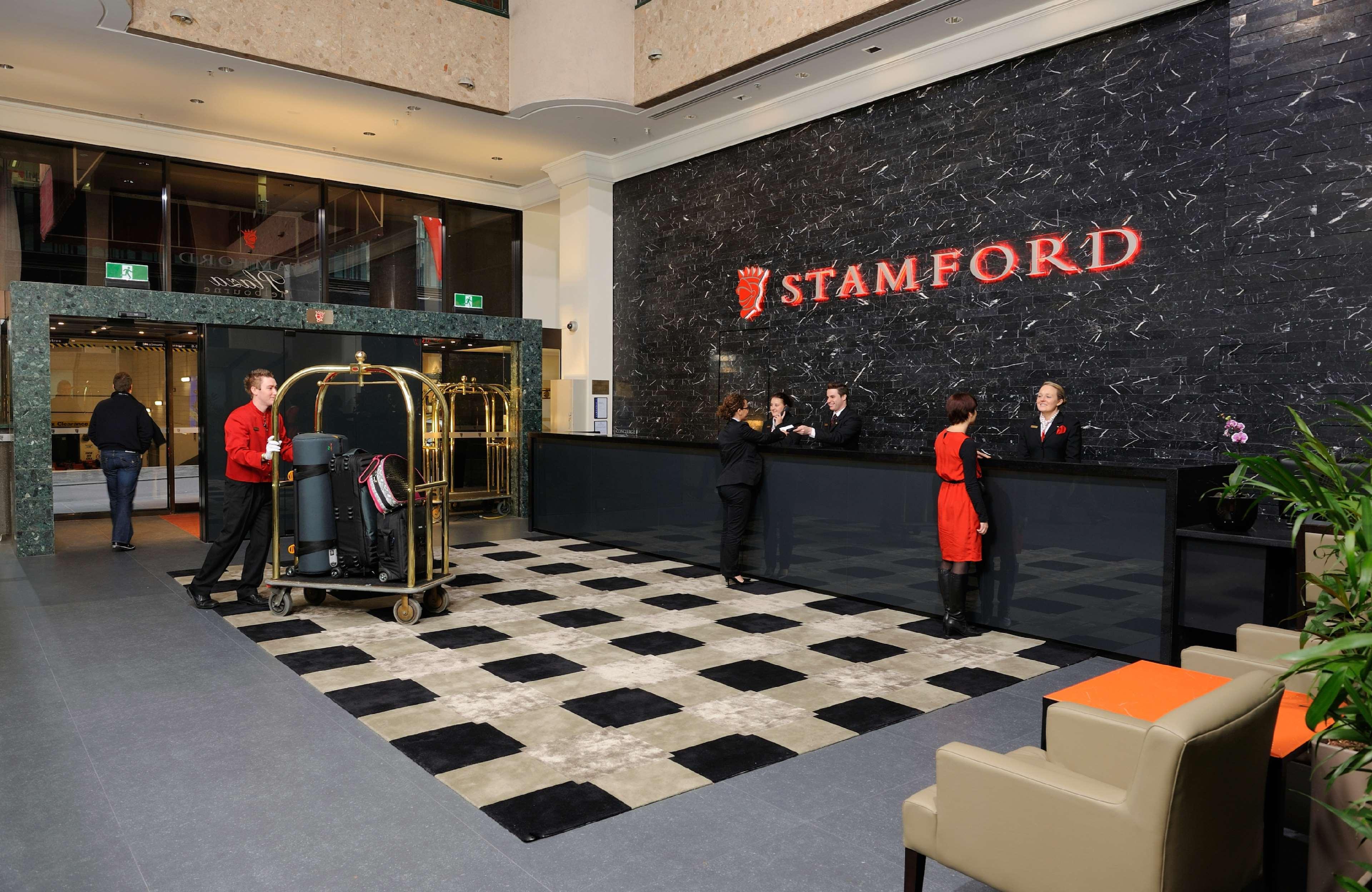 Отель Stamford Plaza Мельбурн Интерьер фото The photo shows the interior of a hotel lobby, specifically the Stamford Hotel. There is a large reception desk where several staff members are attending to guests. In the foreground, a bell cart loaded with luggage is positioned beside the reception