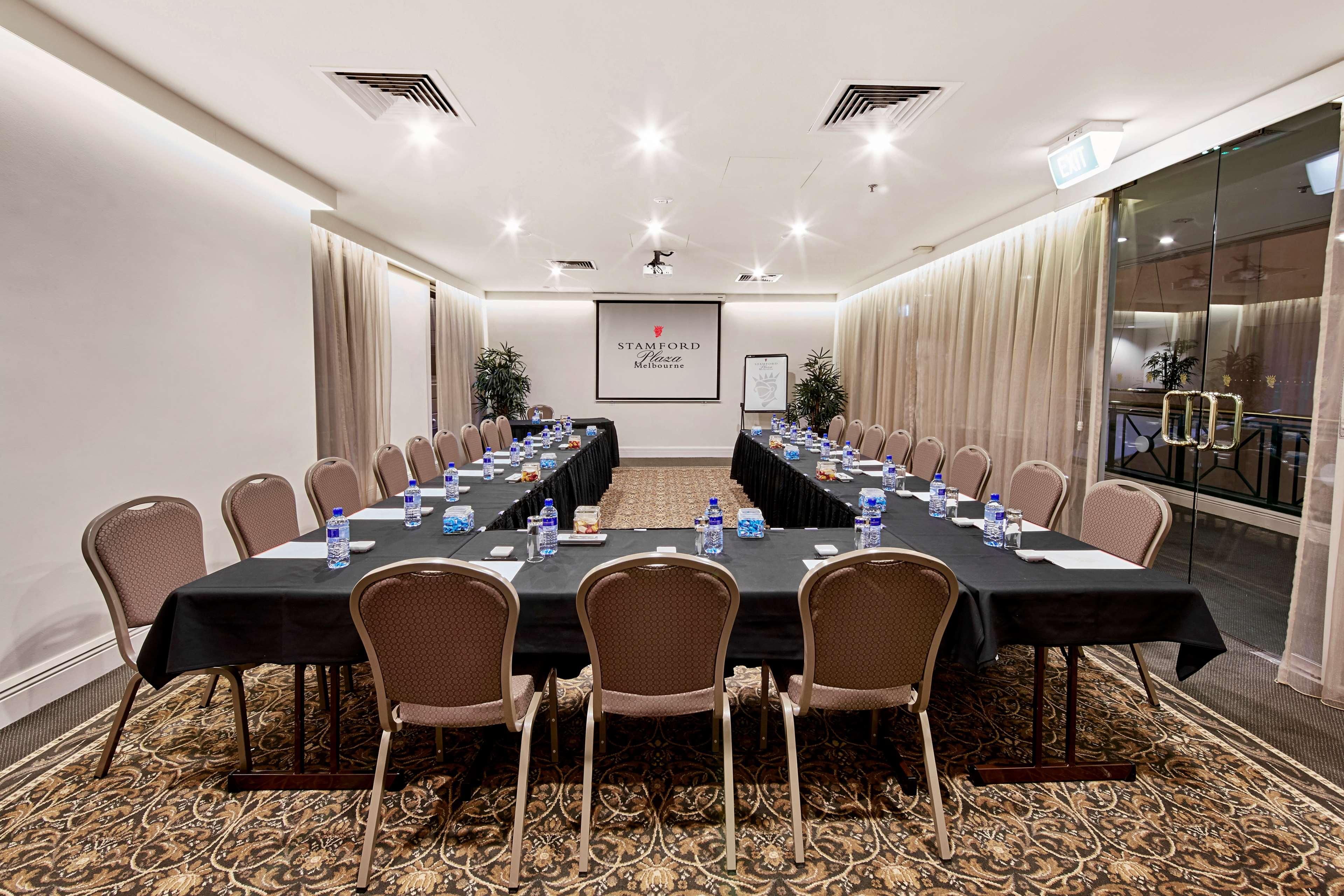 Отель Stamford Plaza Мельбурн Экстерьер фото The photo shows a conference room set up for a meeting. There are several long tables arranged in a U-shape, covered with black tablecloths. Each table has a water bottle and notepad placed at intervals. The room is well-lit, with light-colored walls