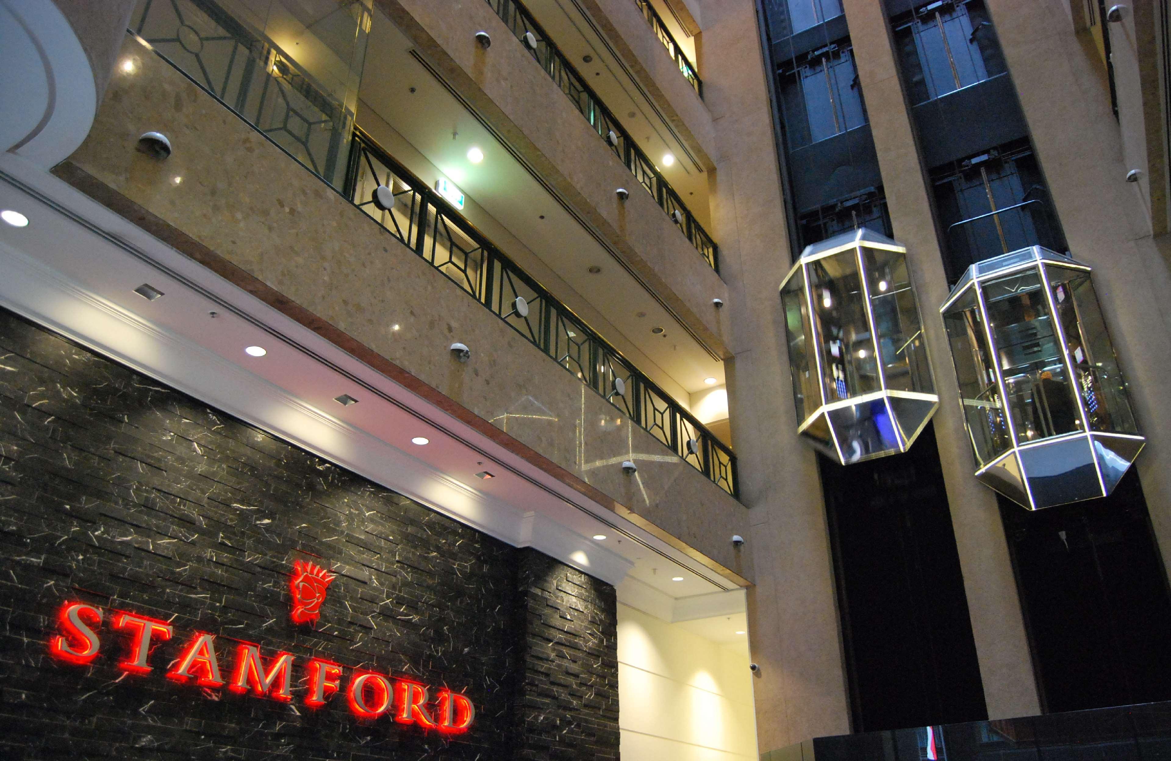 Отель Stamford Plaza Мельбурн Экстерьер фото The photo shows the interior of a building, likely a hotel or commercial space. There is a prominent sign with the name "STAMFORD" in red lettering against a dark stone background. The building features several floors, visible from the photograph, wi