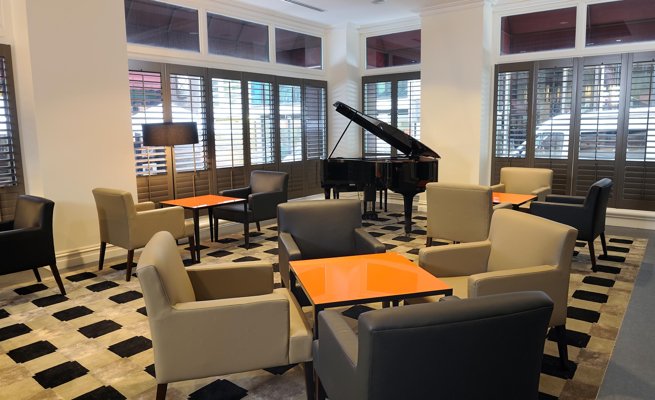 Отель Stamford Plaza Мельбурн Экстерьер фото The photo shows a stylish indoor lounge area. It features several light gray armchairs arranged around orange square tables. In the corner, there is a black grand piano, adding an elegant touch to the space. The room has large windows with wooden shu