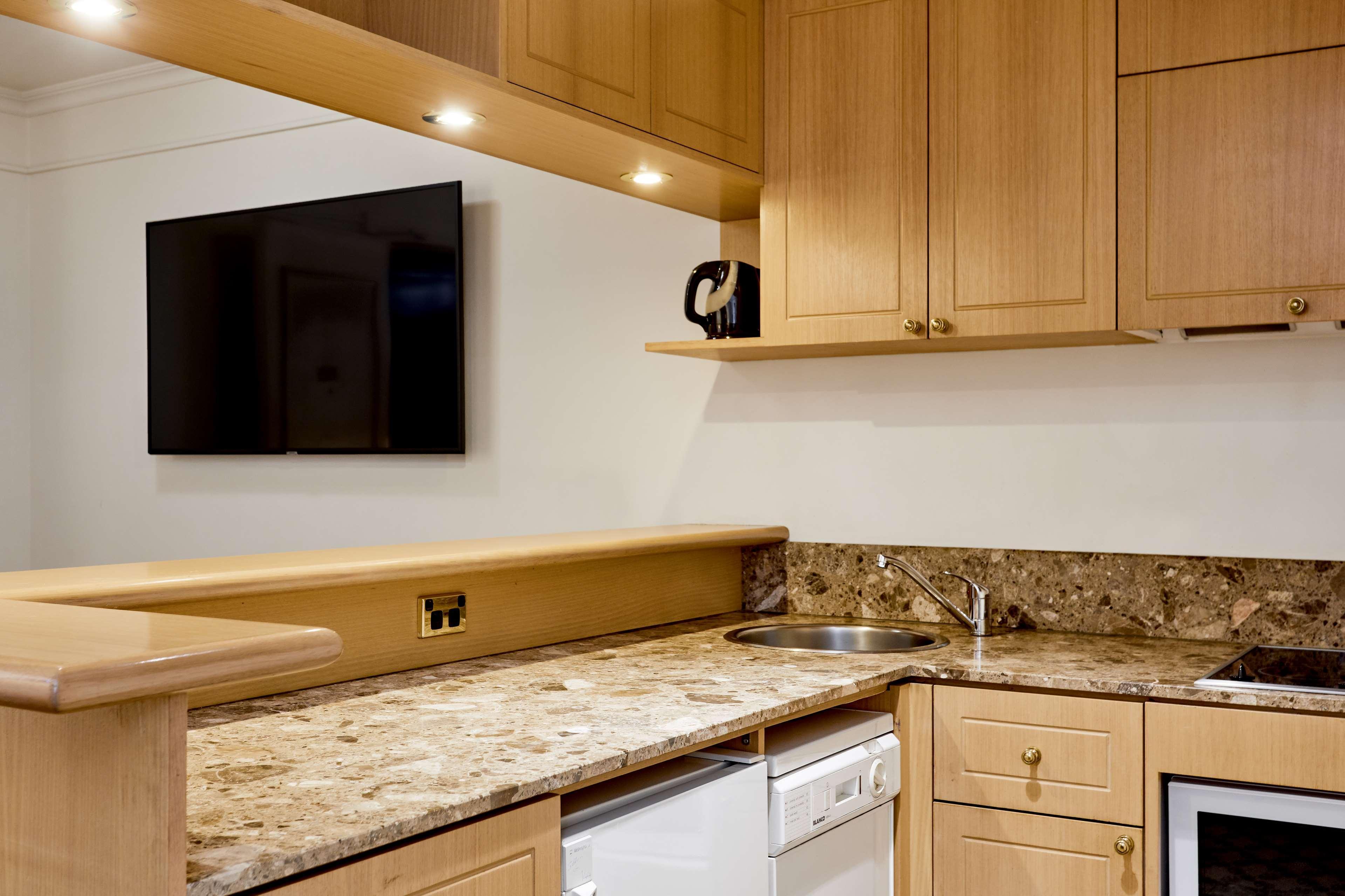 Отель Stamford Plaza Мельбурн Экстерьер фото The photo shows a modern kitchen with wooden cabinetry and a granite countertop. There's a stainless steel sink integrated into the countertop, and appliances including a dishwasher and possibly a coffee maker on the shelf above. A flat-screen televi