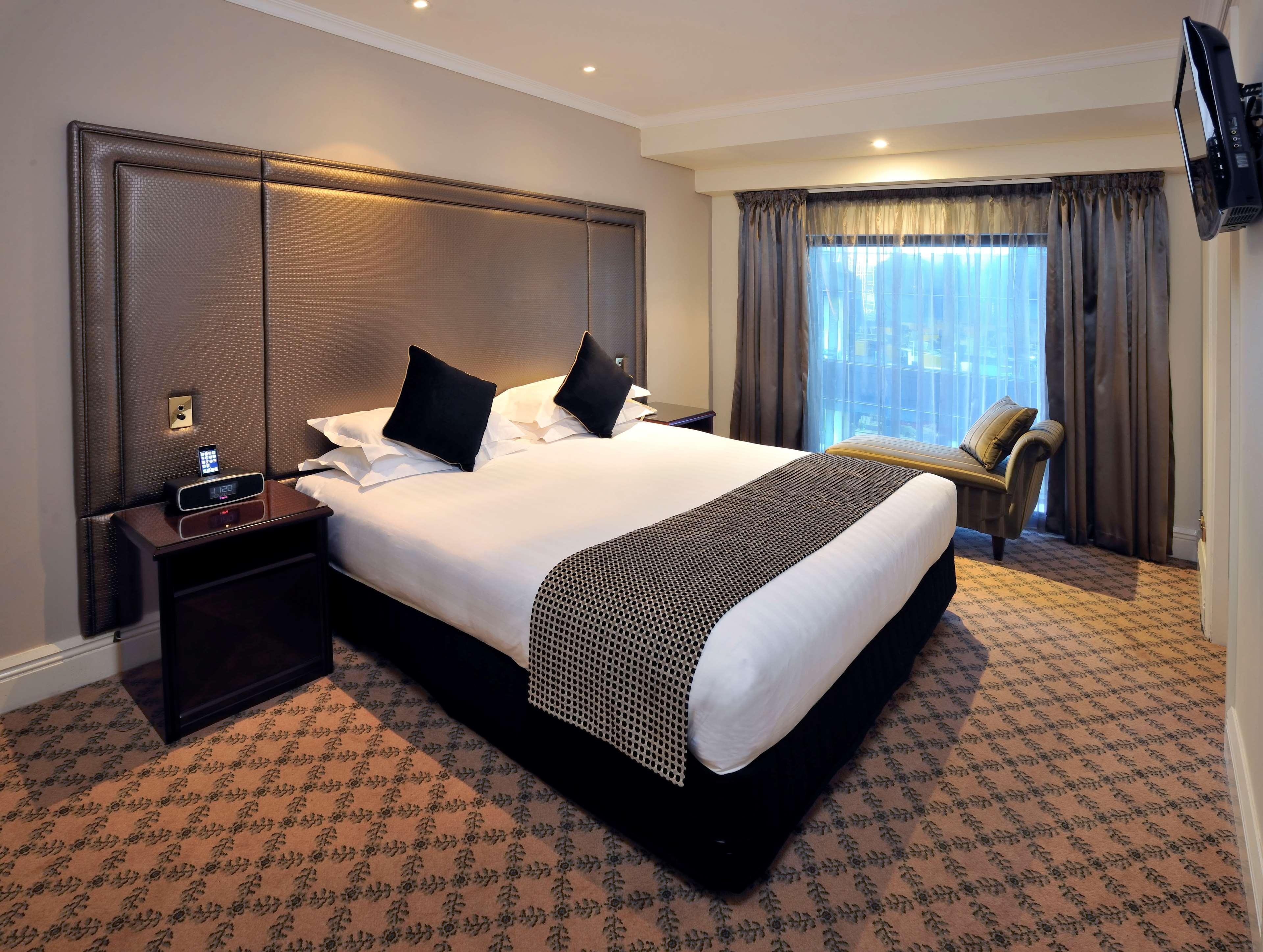 Отель Stamford Plaza Мельбурн Экстерьер фото The photo shows a hotel room featuring a neatly made king-sized bed with dark pillows. The headboard is upholstered in a dark material, and there is a decorative blanket at the foot of the bed. Beside the bed are two nightstands, each equipped with l