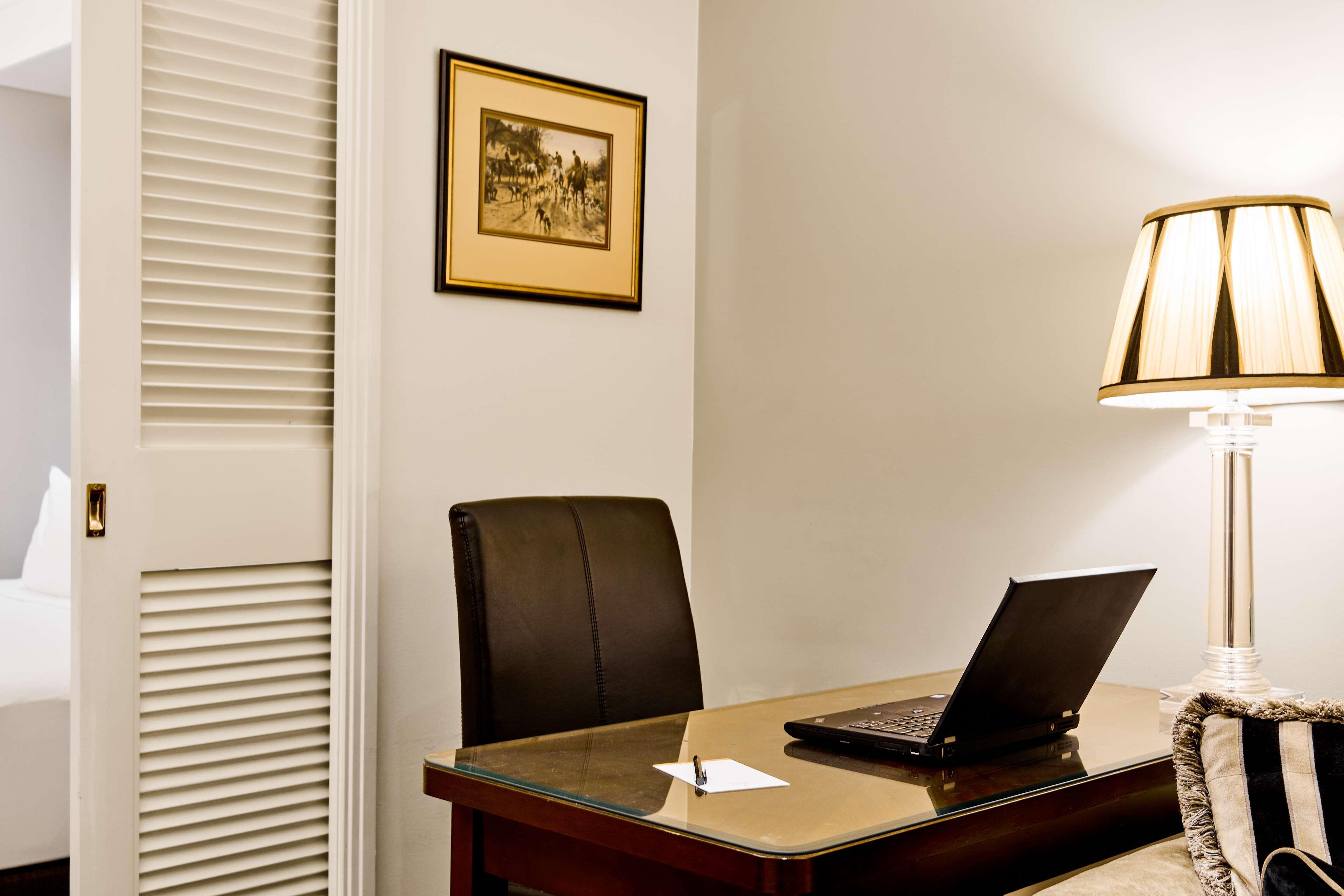 Отель Stamford Plaza Мельбурн Экстерьер фото The photo shows a small office or workspace. There is a desk with a glass surface, and on it sits a laptop computer. A piece of paper or notecard is also placed on the desk. Behind the desk is a black office chair. A wall lamp with a striped shade is