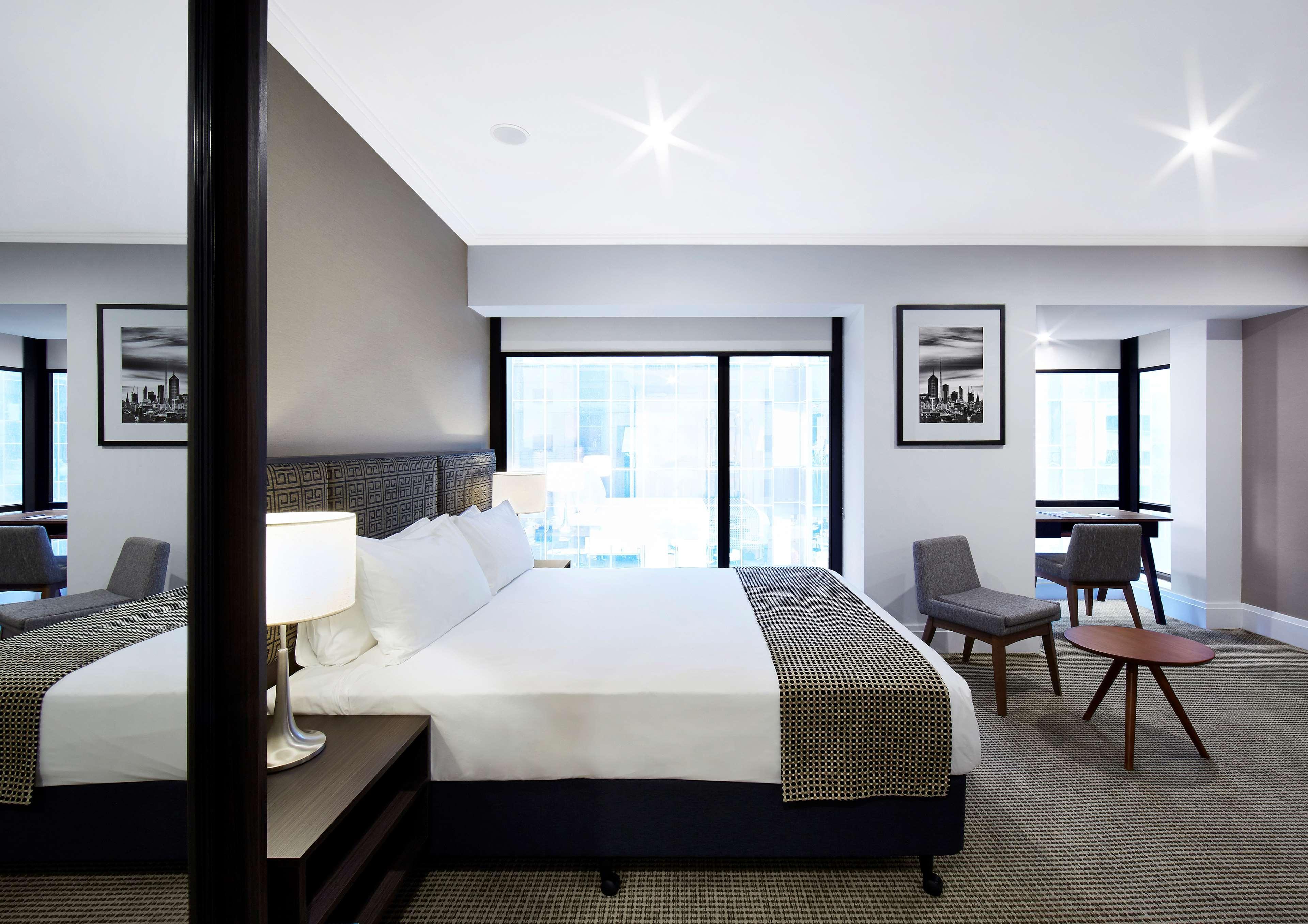 Отель Stamford Plaza Мельбурн Экстерьер фото The image shows a modern hotel room featuring a large bed with white linens and a gray throw blanket. There are two windows that allow natural light to enter the space, and the walls are painted in a light color. In addition to the bed, there are two