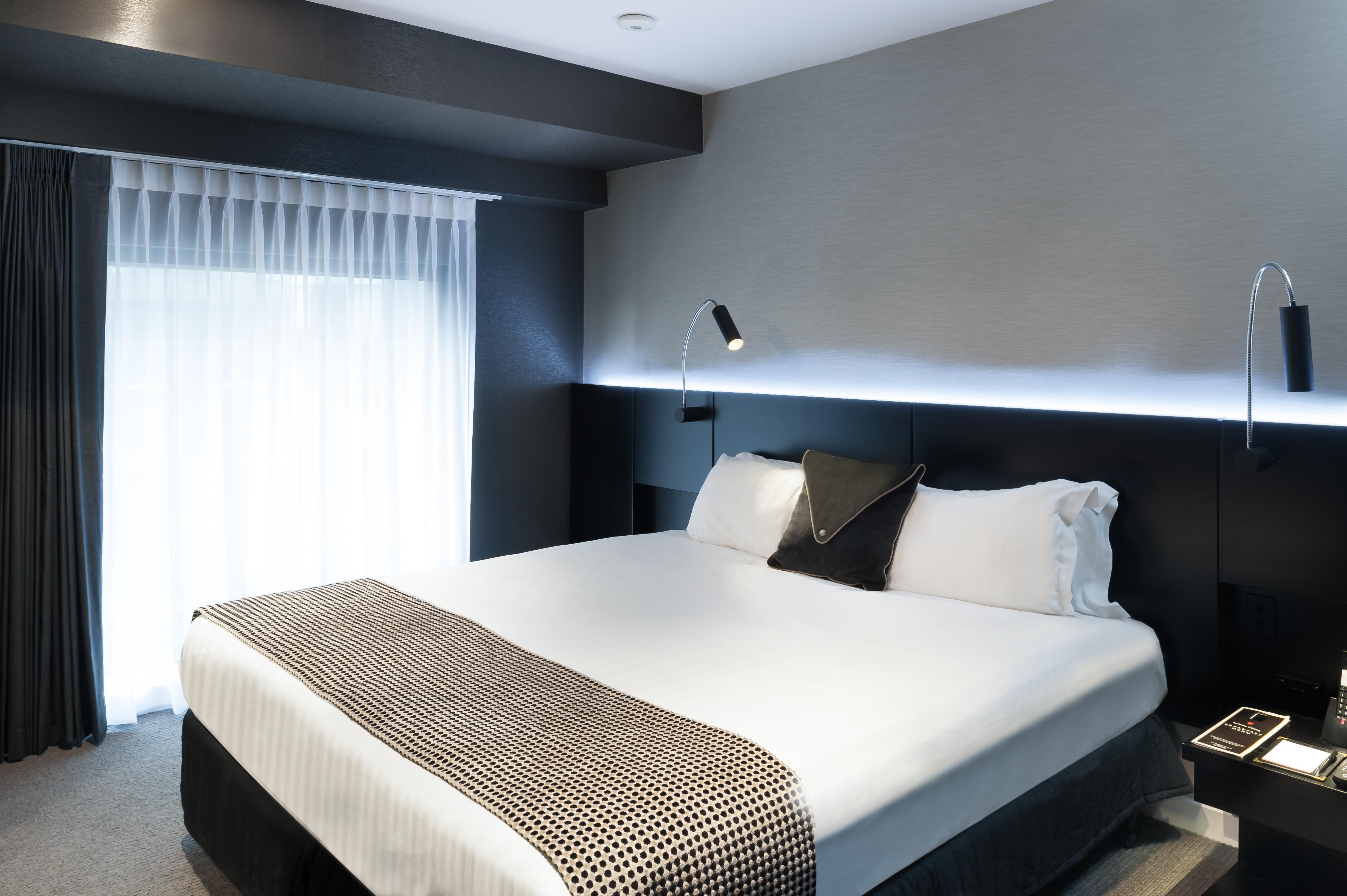 Отель Stamford Plaza Мельбурн Экстерьер фото The photo shows a modern hotel room featuring a large bed with white linens and a decorative throw blanket. The room has a sleek and contemporary design, with gray walls and a soft lighting strip along the headboard. A window is adorned with sheer cu