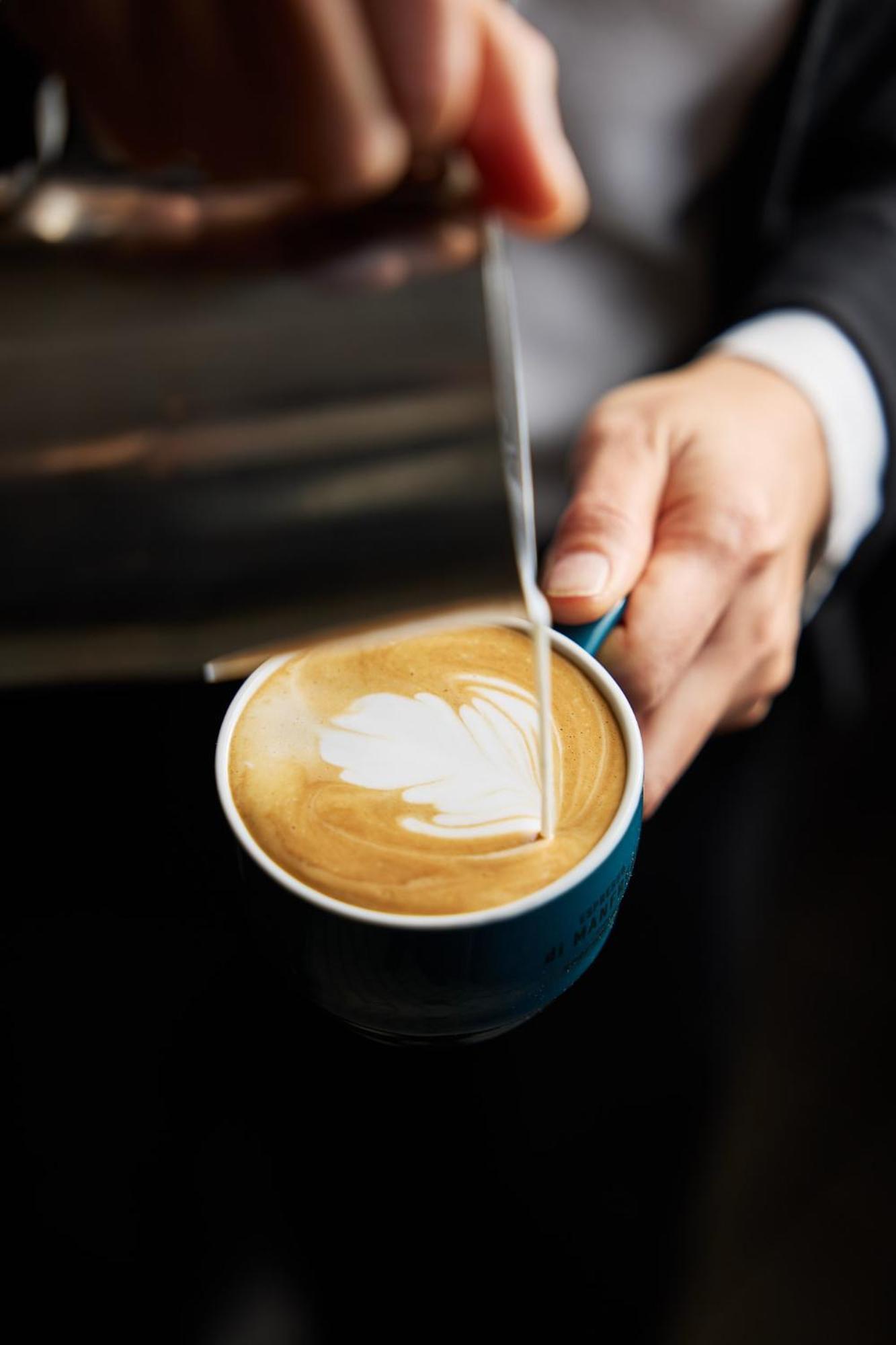 Отель Stamford Plaza Мельбурн Экстерьер фото The photo shows a close-up of a hand holding a cup of coffee with a latte art design on the surface. The coffee appears to be a creamy, caramel color, likely a latte or cappuccino. A small stream of milk is being poured into the cup, creating a heart
