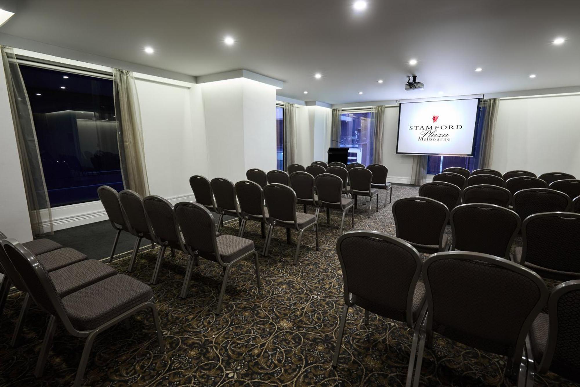 Отель Stamford Plaza Мельбурн Экстерьер фото The image shows a conference or meeting room set up for an event. There are rows of gray chairs arranged facing a large screen displaying the name "Warford House." The room is well-lit with overhead lighting and has large windows covered with light-c