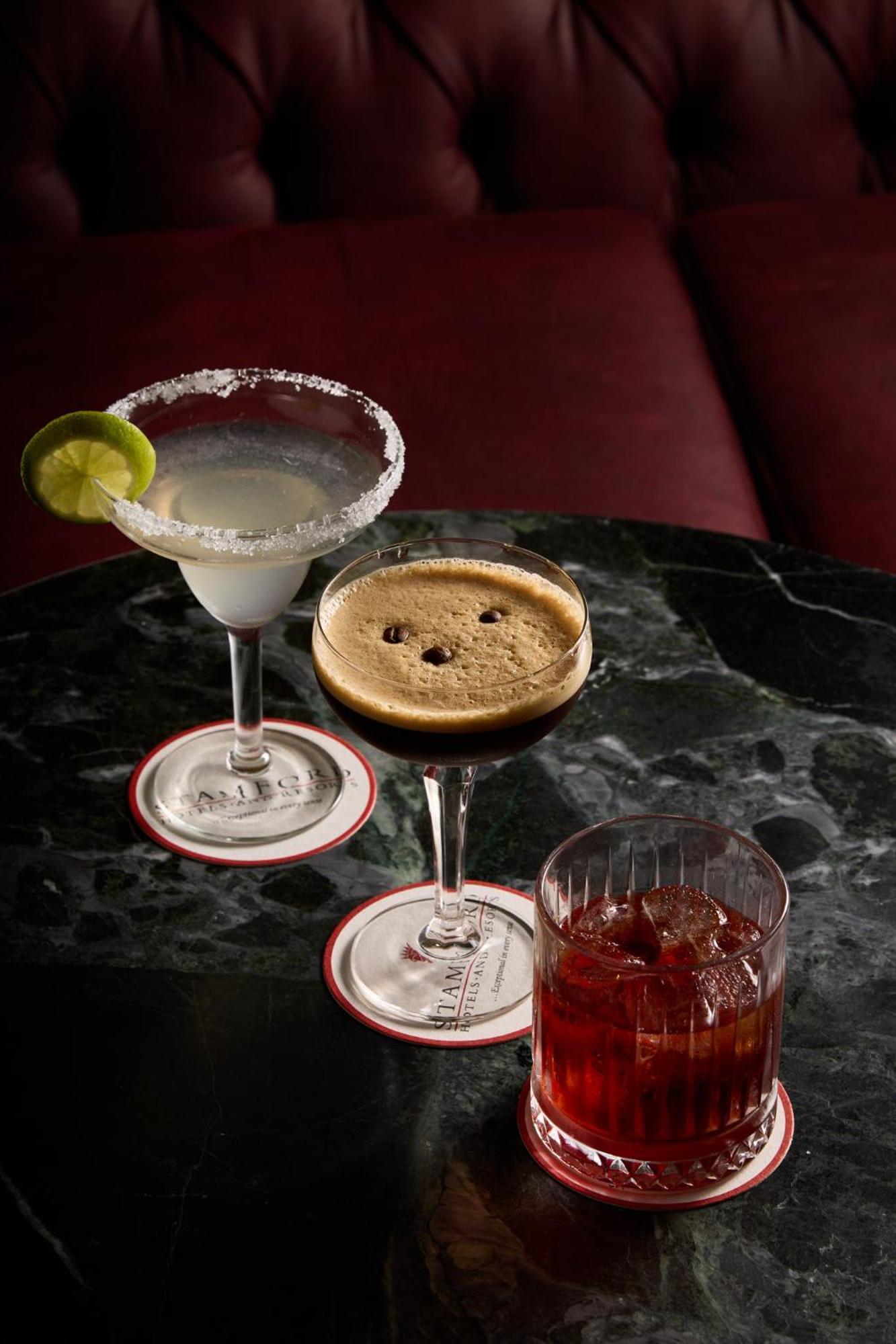 Отель Stamford Plaza Мельбурн Экстерьер фото The photo displays three different cocktails arranged on a dark marble table. 

1. On the left, there is a margarita glass filled with a clear drink, garnished with a lime slice and featuring a salted rim.
2. In the center, a coupe glass holds a dark