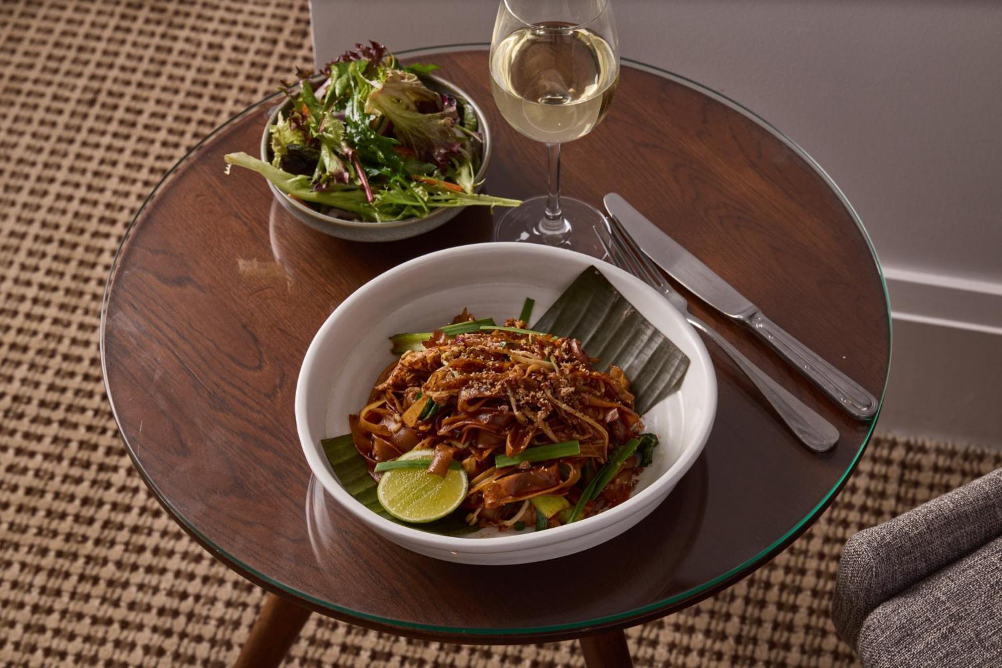 Отель Stamford Plaza Мельбурн Экстерьер фото The photo shows a meal served on a wooden table. In the foreground, there is a bowl of stir-fried noodles garnished with green onions and a lime wedge, set atop a banana leaf. Next to it, there is a bowl of mixed green salad. A glass of white wine si