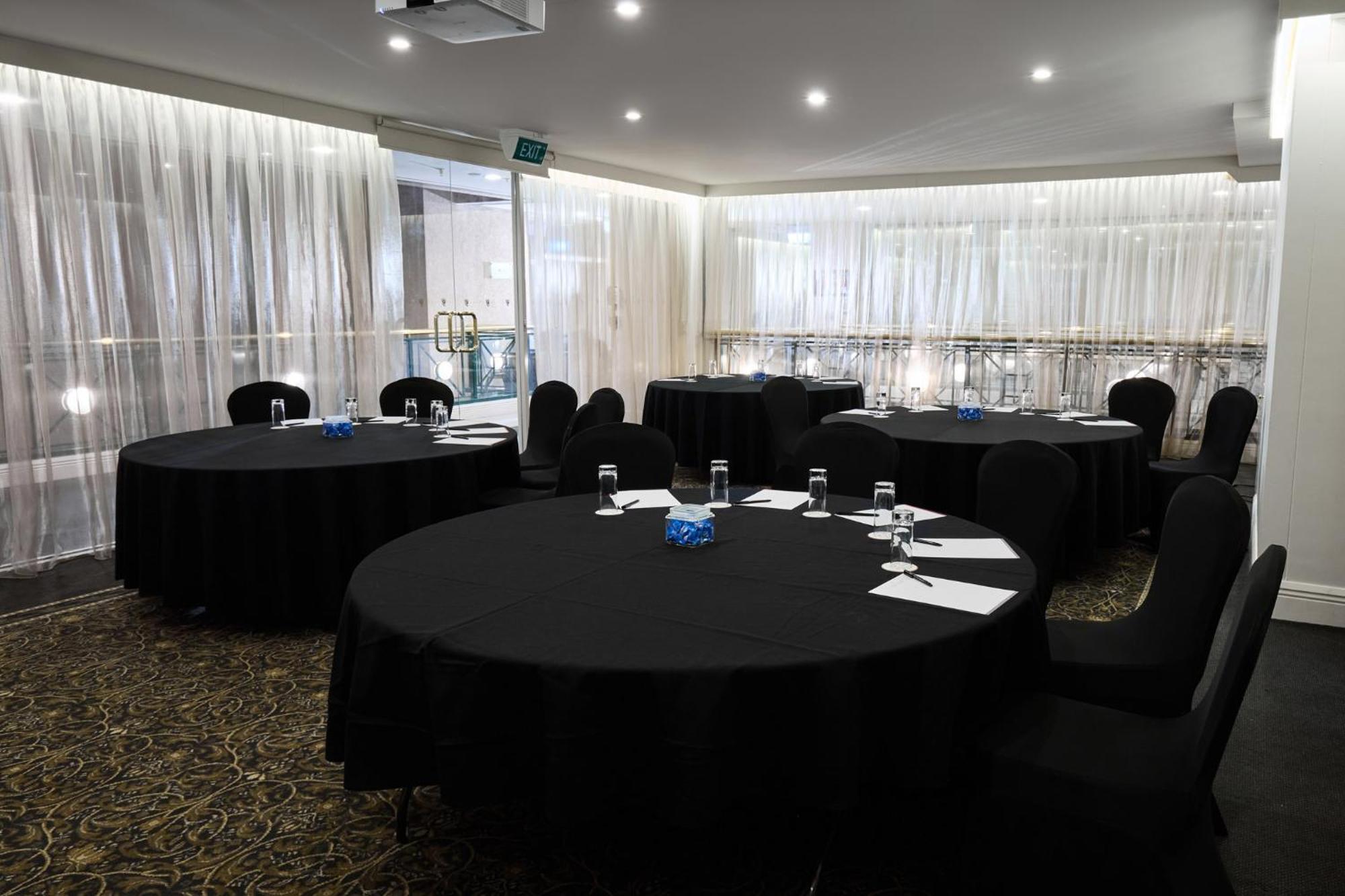 Отель Stamford Plaza Мельбурн Экстерьер фото The photo shows a meeting or conference room set up with several round tables covered in black tablecloths. Each table is accompanied by black chairs. There are some white notepads and pens placed on the tables, and a small decorative item, possibly 
